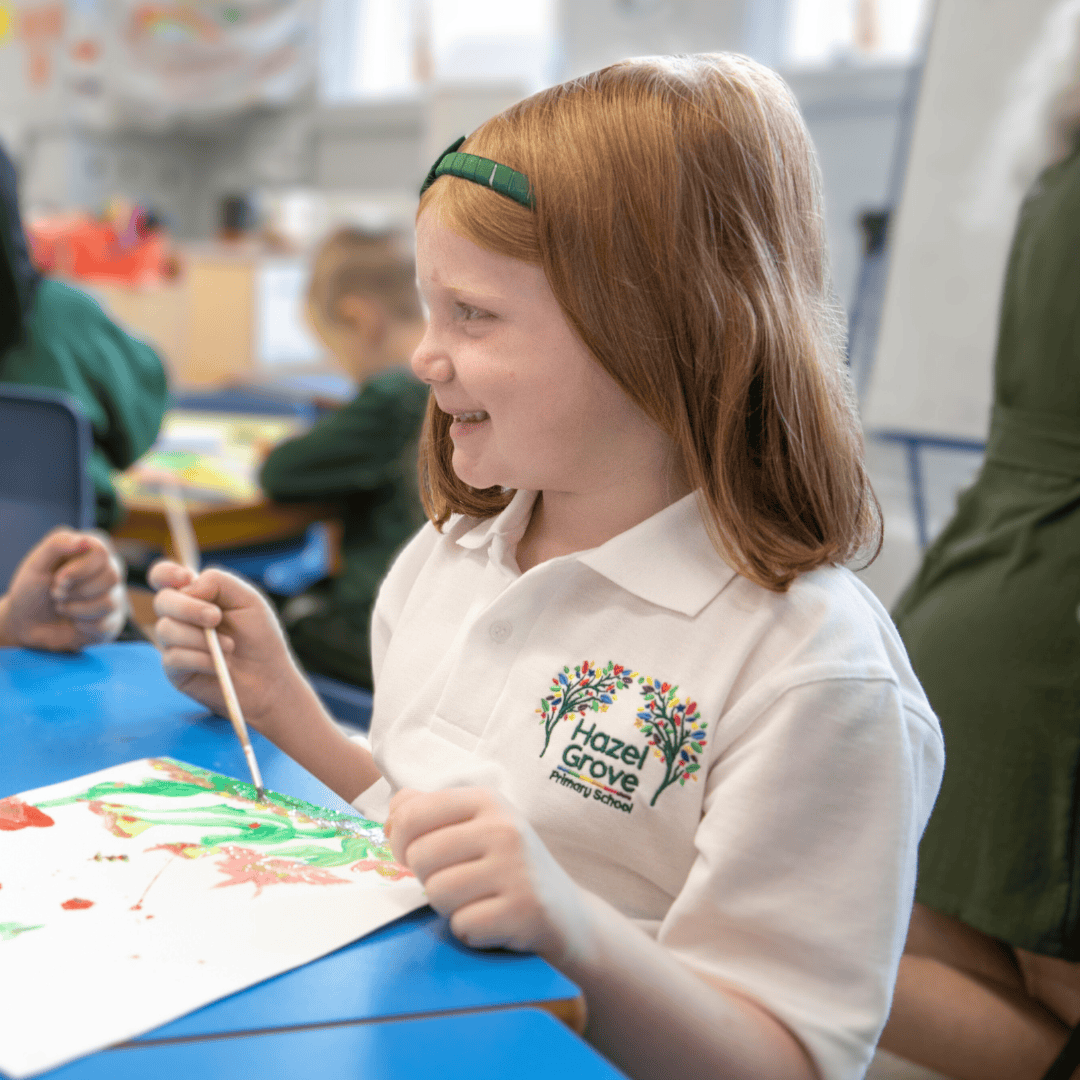 A girl wears the Hazel Grove Pre-School uniform of white polo with school logo while painting in a classroom.