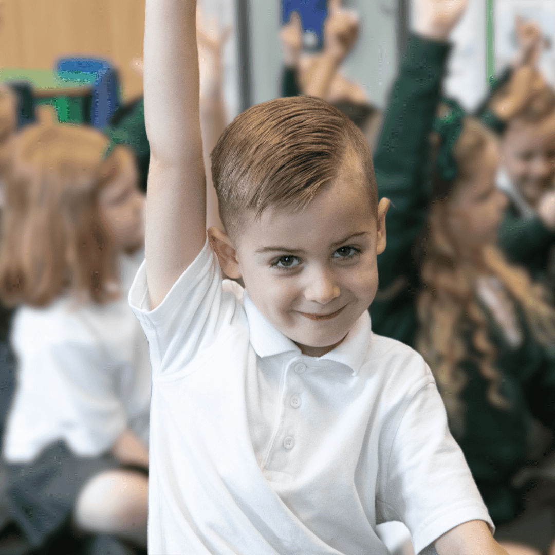 A boy wears the Hazel Grove Pre-School uniform of white polo shirt and raises his hand in classroom.